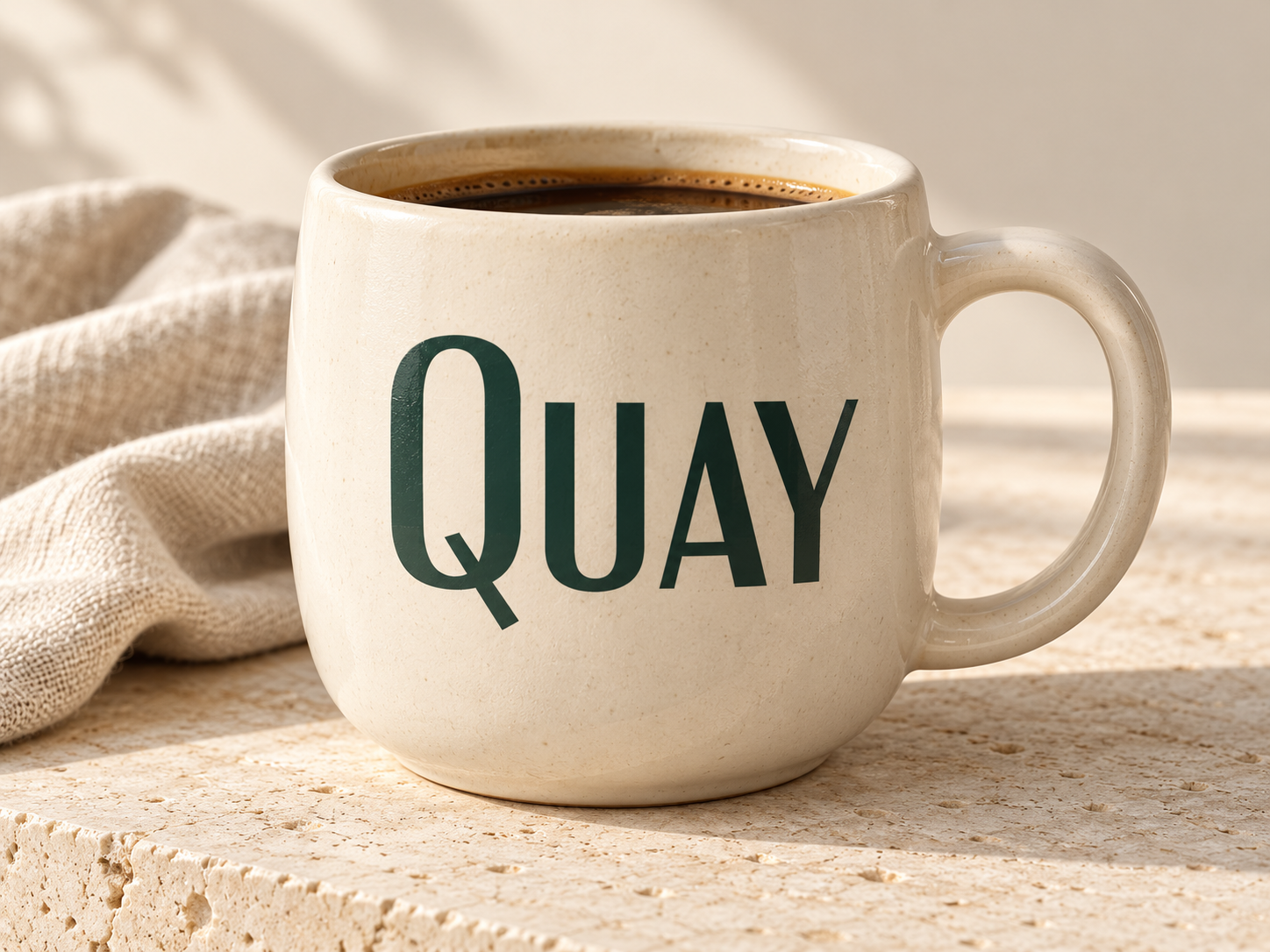 Generated concept of a cream ceramic coffee mug with dark green Quay lettering on a warm stone surface.